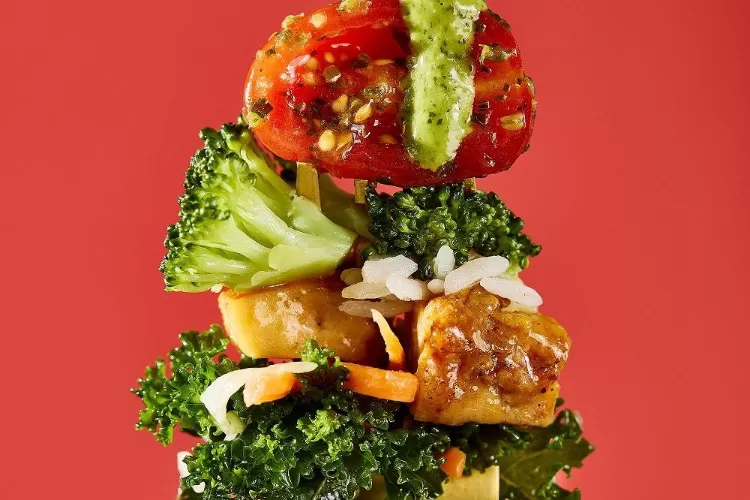 Bolay Fresh Bold Kitchen ingredients stack up, including pesto, tomatoes, broccoli, rice, teriyaki chicken, and kale on a red background